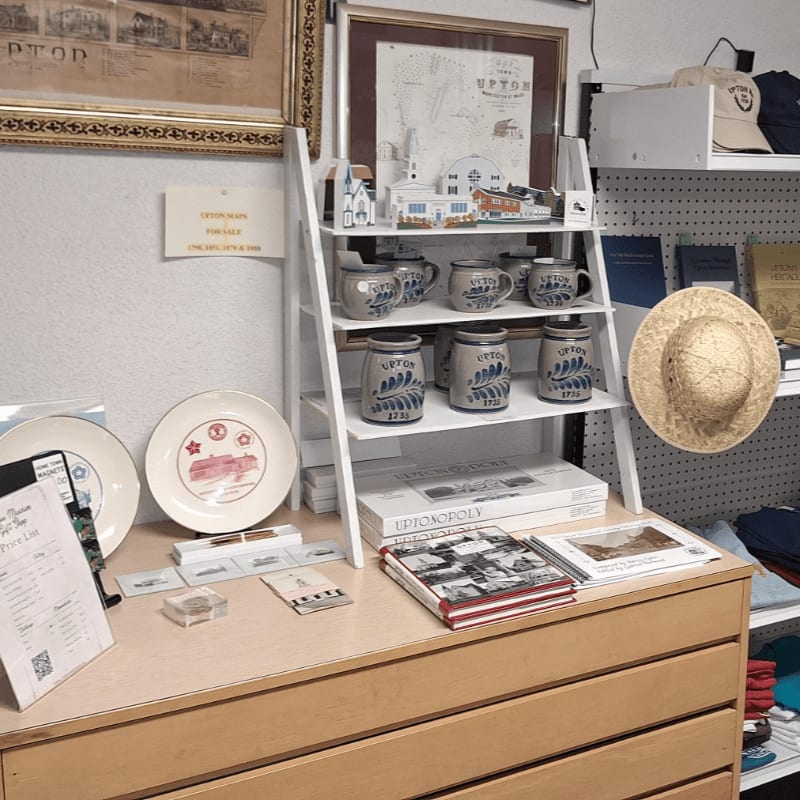 In the foreground, a wooden counter displays plates, books, and mugs, while the background features shelves stocked with more merchandise and a framed map.