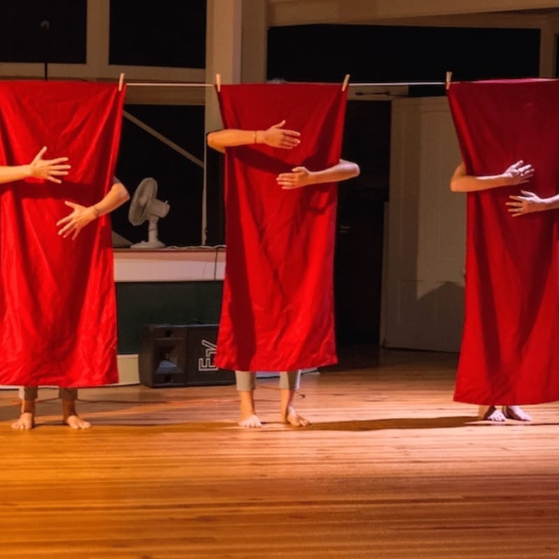 Three figures, draped in red fabric and holding their hands to their chests, stand on a wooden stage with a fan and speaker visible in the dimly lit background.