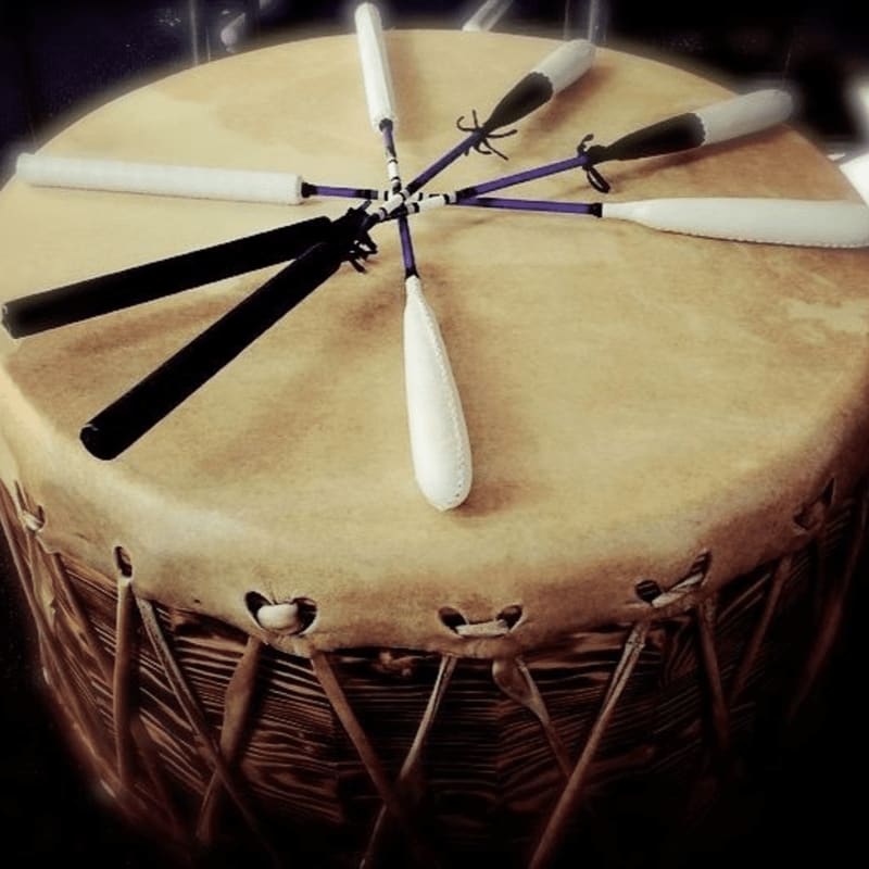 A large, tan drum with lacing around its edge is topped with a fan of drumsticks, some white and some black, against a dark background.
