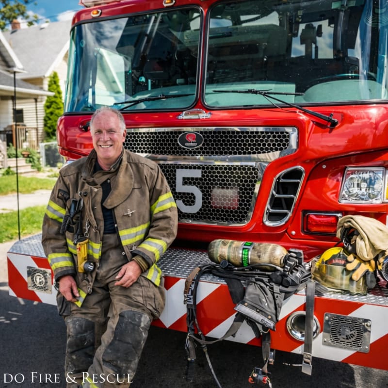 A smiling firefighter in full gear sits on the front bumper of a red fire truck, with a residential street and houses visible in the background.