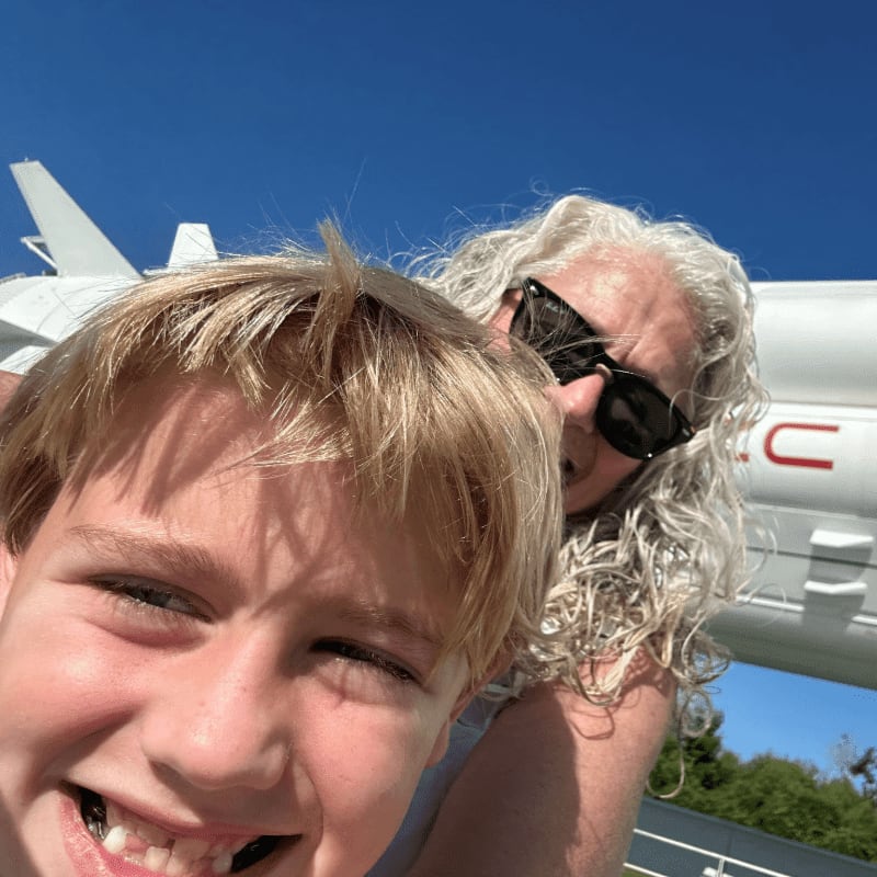 A young boy with blond hair smiles in the foreground, while a woman with curly gray hair and sunglasses poses behind him with a white airplane in the background.