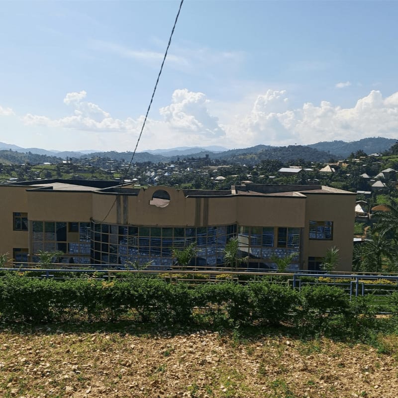A modern tan building with large windows sits in the foreground, backed by a lush green hillside dotted with houses and distant mountains under a cloudy blue sky.