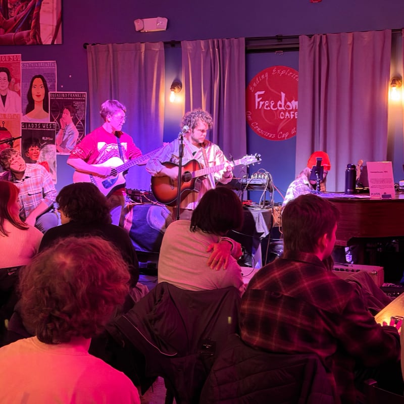 Two musicians play acoustic guitars on a stage in front of a seated audience, with posters and a cafe sign visible in the background.