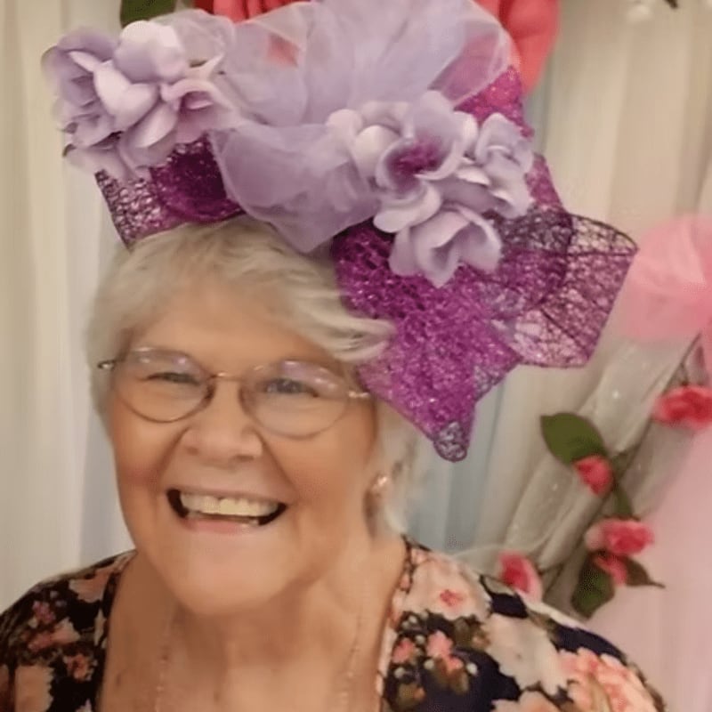 A smiling elderly woman in a floral top wears a large, ornate purple and lavender hat, with a soft pink and white draped background adorned with artificial flowers.