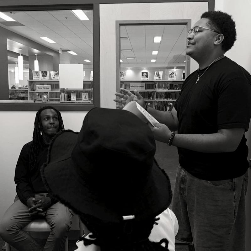 A man with glasses and a black t-shirt gestures with a book while speaking to an audience in a library, with bookshelves visible in the background.