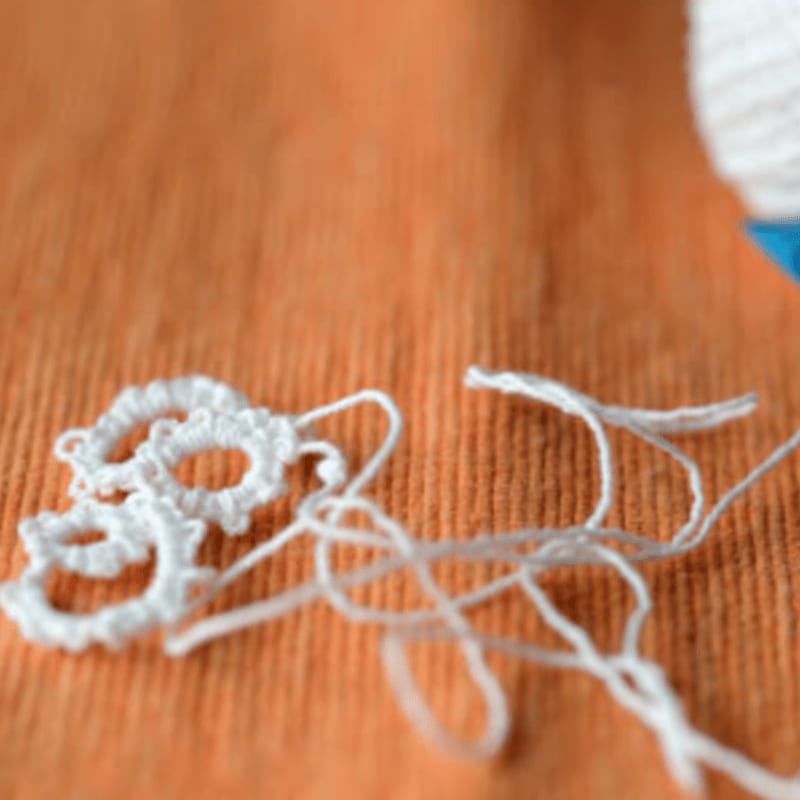Delicate white tatting lace lies tangled with thread in the foreground, against a blurred orange background with a hint of a white yarn ball.