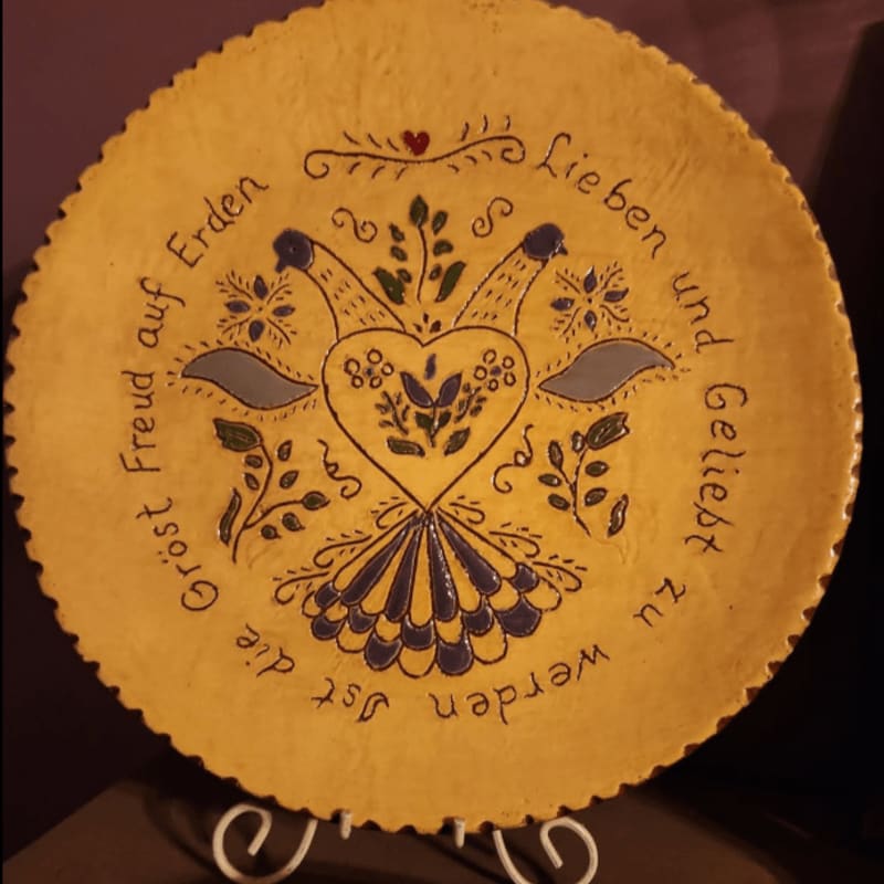 A yellow decorative plate with folk art birds and a heart is displayed on a white stand against a dark background.