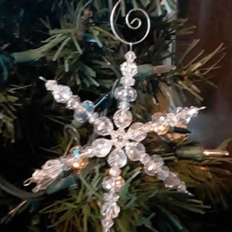 A clear beaded snowflake ornament hangs on a green Christmas tree with blurred lights in the background.