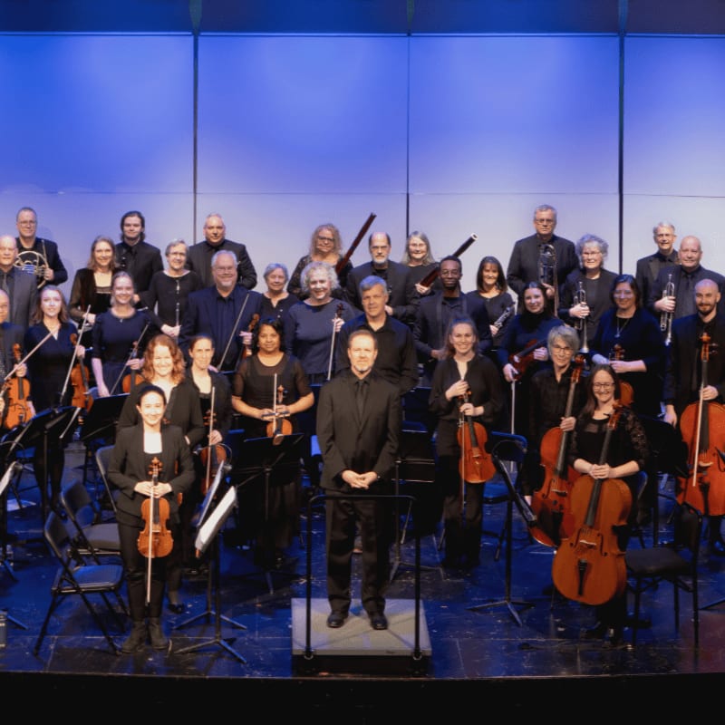 A full orchestra stands on a stage with a blue backdrop, musicians in formal attire holding their instruments.