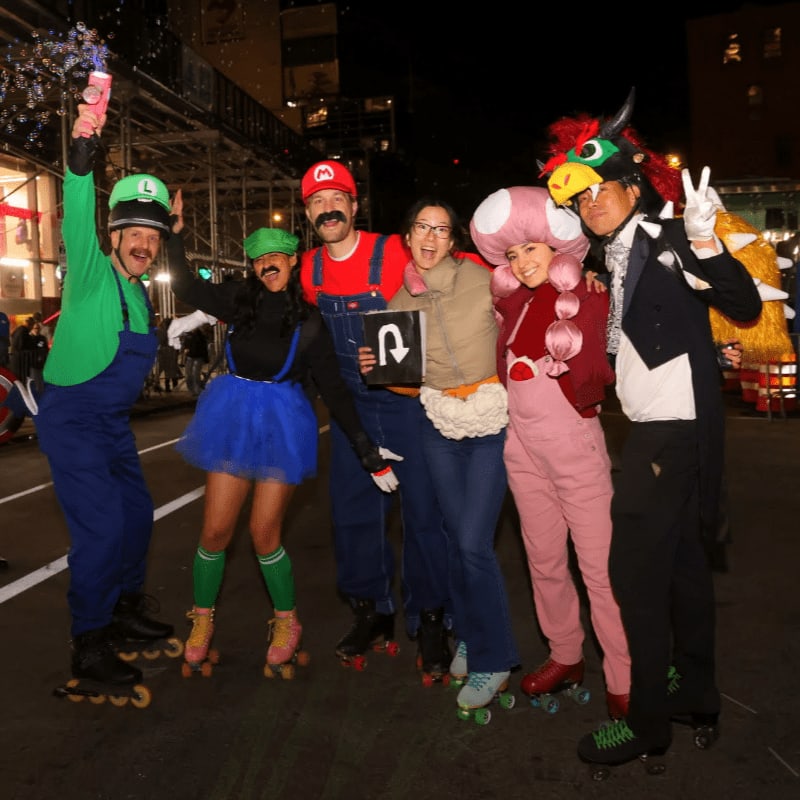 Six people in Mario-themed costumes, some on roller skates, pose in front of a nighttime street scene with buildings.