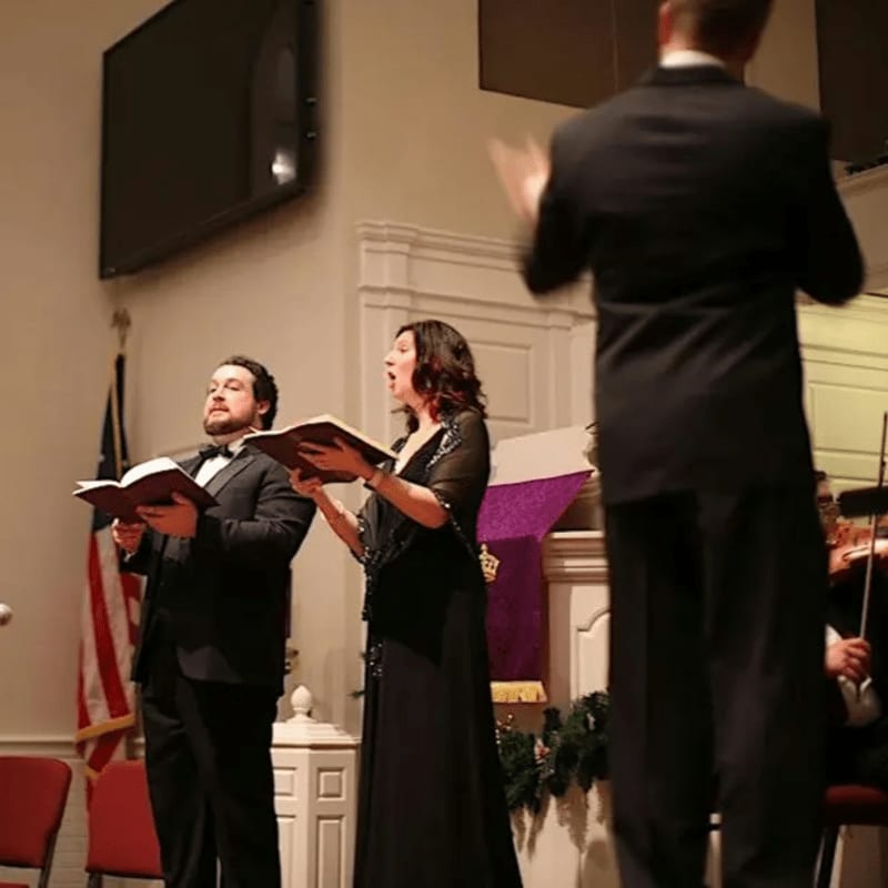 Two singers in formal attire hold open books in the foreground, while a conductor stands with his back to the camera in front of a decorated altar and an American flag in the background.