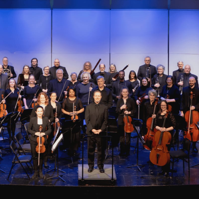 A full orchestra stands on a stage with a blue backdrop, with musicians in the foreground holding their instruments.