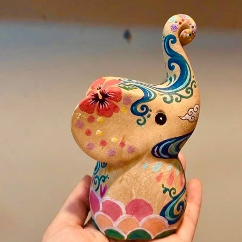 A hand holds a small, vibrantly painted ceramic elephant with floral and wave motifs against a softly blurred background.