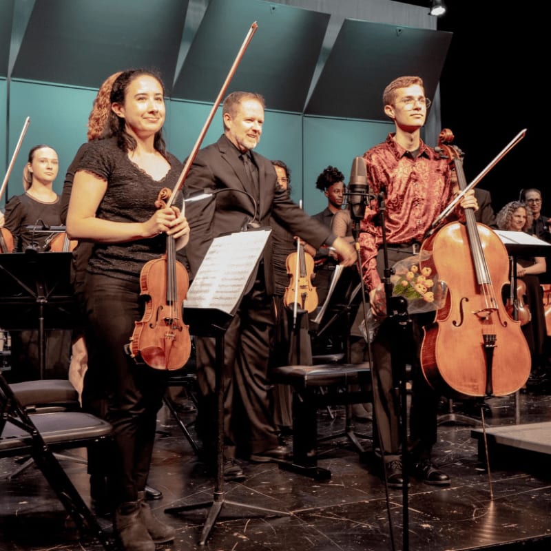 A woman with a violin and a young man with a cello stand in the foreground, while an orchestra plays behind them on a stage.
