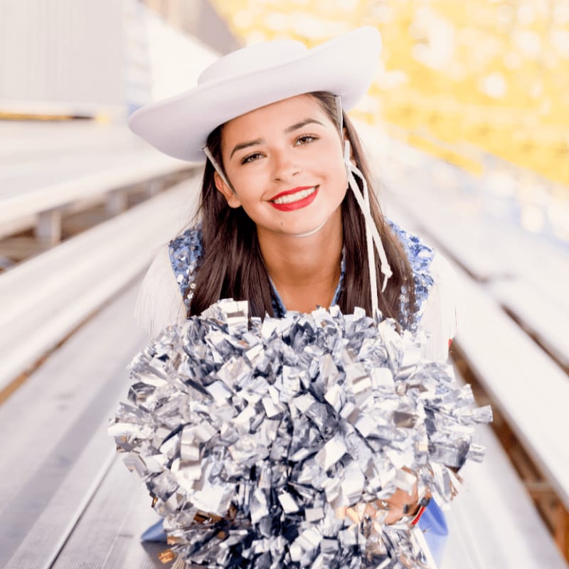 A smiling cheerleader in a white cowboy hat and blue sequined vest holds a silver pom-pom in the foreground, while blurred bleachers and yellow foliage form the background.