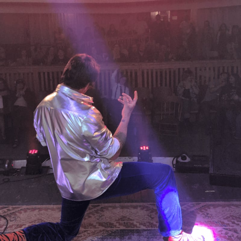 A performer in a shiny shirt and blue pants kneels on a patterned rug in the foreground, while an audience watches from the background.