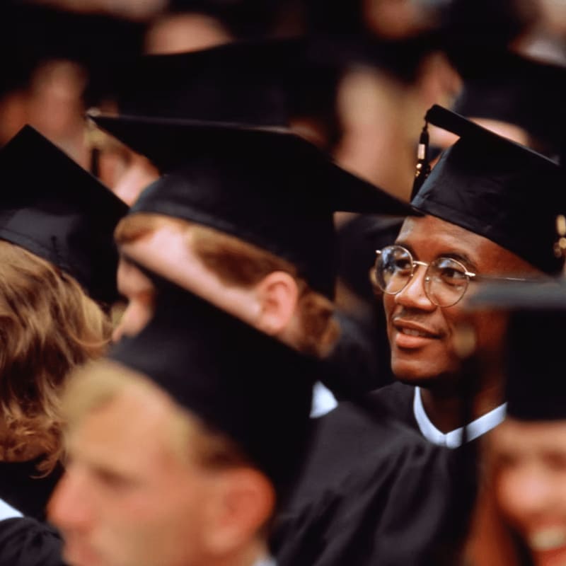A group of graduates in black caps and gowns are seated, with a man in the foreground looking to the side with a smile.