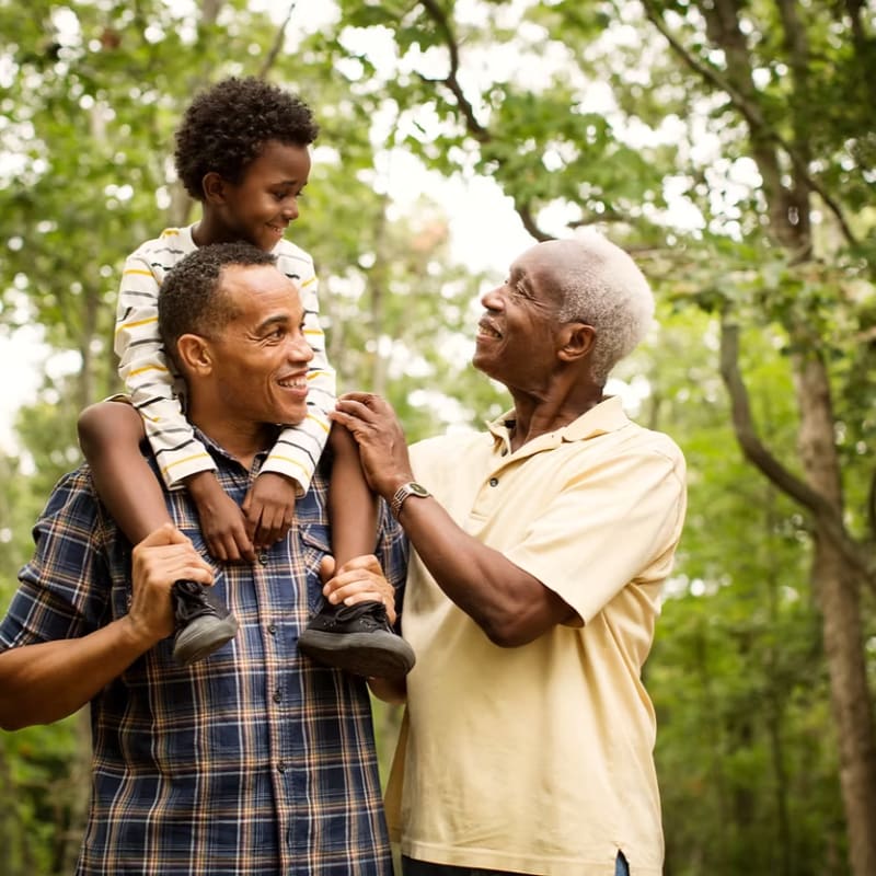 A smiling young boy sits on his father's shoulders, looking at his grandfather, all surrounded by lush green trees.