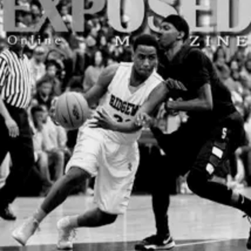 A basketball player in a white jersey dribbles the ball while a player in a black jersey defends, with a blurred crowd in the background.