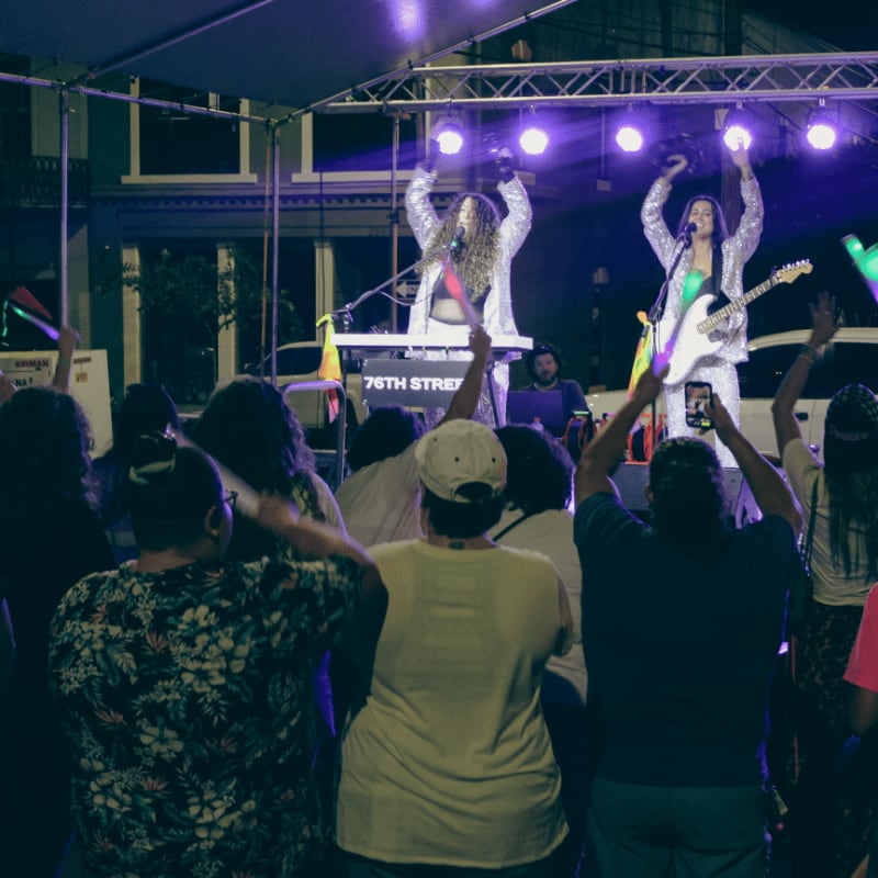A crowd with raised hands enjoys a live band performing on a brightly lit stage with buildings in the background.