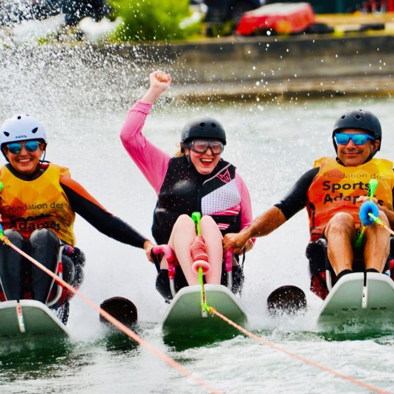 Three smiling individuals in adaptive water skis, with one raising a fist in triumph, are pulled across a lake with a blurred background of green trees and a red object.