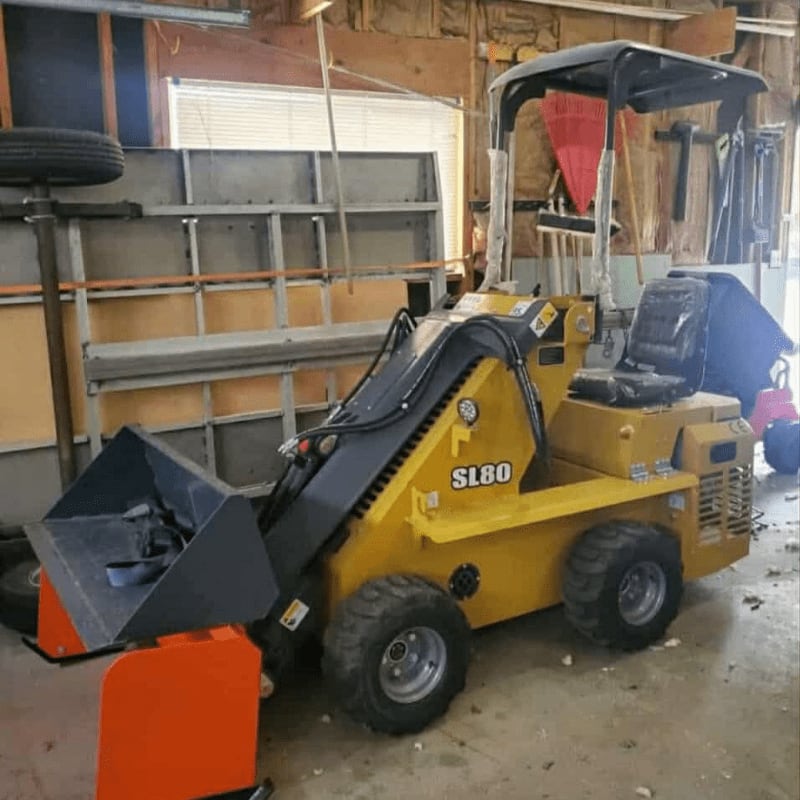 A yellow skid steer loader with an orange attachment sits in the foreground of a cluttered garage with shelves and tools in the background.