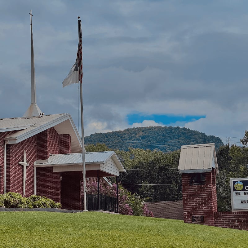 A brick church with a steeple and an American flag in the foreground stands against a backdrop of a tree-covered mountain under a cloudy sky.