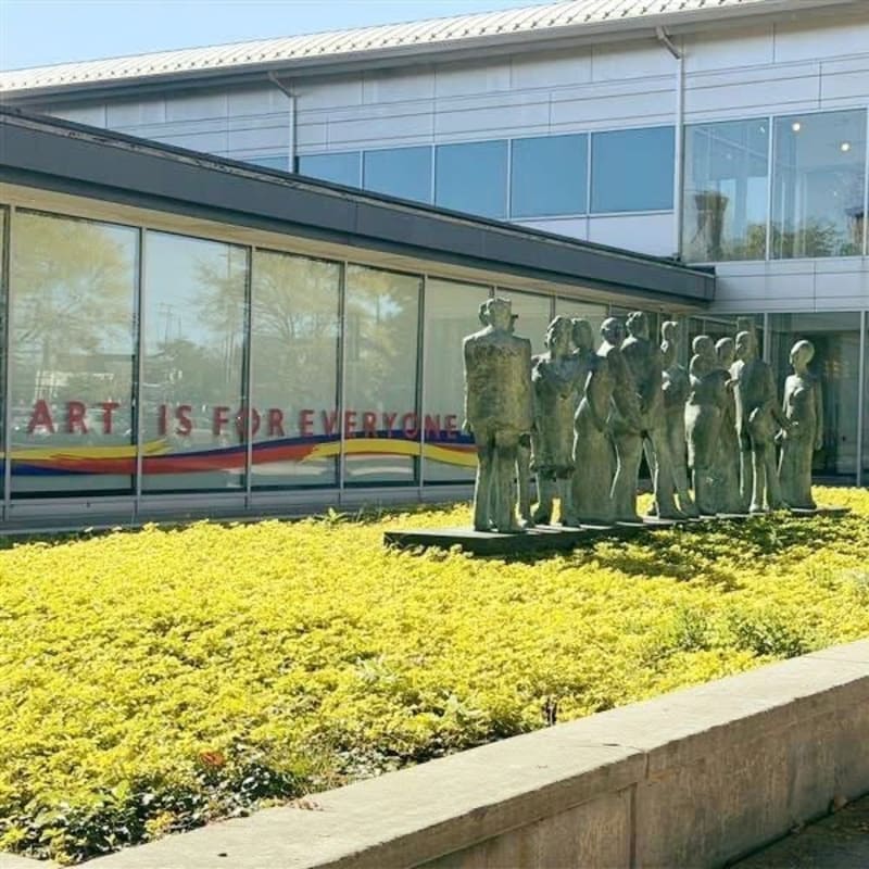 A group of abstract sculptures stands in a vibrant yellow garden in front of a modern building with large windows displaying the words "ART IS FOR EVERYONE."