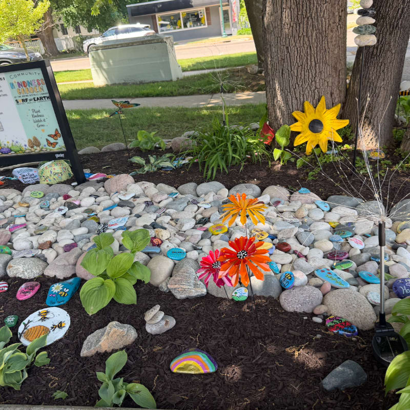Colorful painted rocks and flowers adorn a garden bed in the foreground, with a tree and a street visible in the background.
