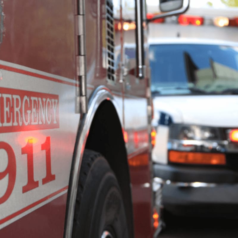 A close-up of a red emergency vehicle with "911" visible in the foreground, and a blurred white ambulance in the background.