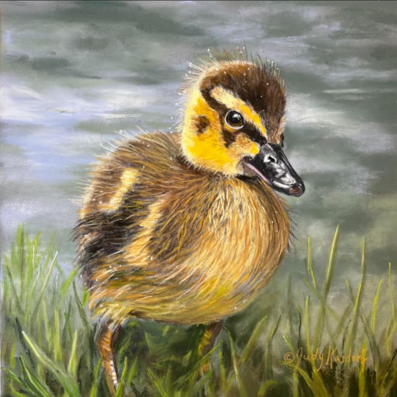 A fluffy duckling stands in lush green grass with a blurred, watery background.