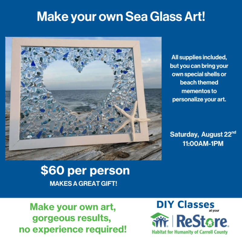 A framed sea glass heart art piece with a starfish sits on a wooden surface in the foreground, with a blurred ocean and sky in the background.