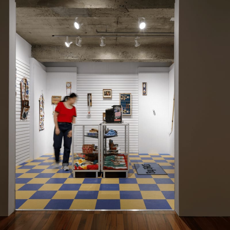 A woman in a red shirt walks past display cases filled with items on a checkered blue and yellow floor, with slatted walls and track lighting in the background.