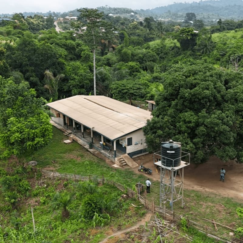 A low-rise building with a water tower sits in the foreground, surrounded by lush green trees and rolling hills in the background.