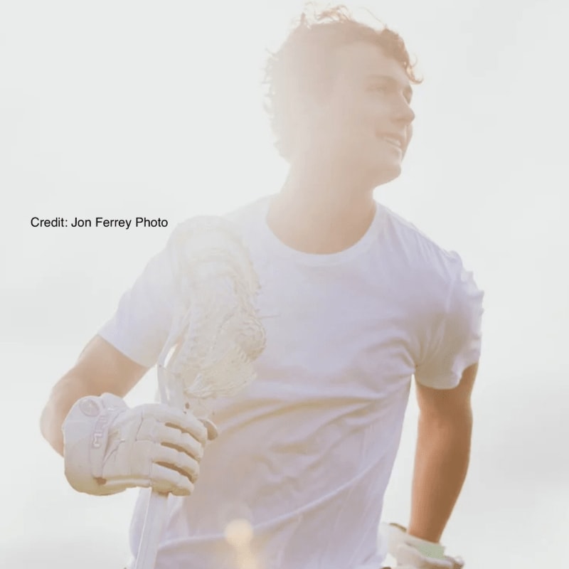 A young man in a white t-shirt and gloves holds a golf club in the foreground, with a bright, hazy sky in the background.