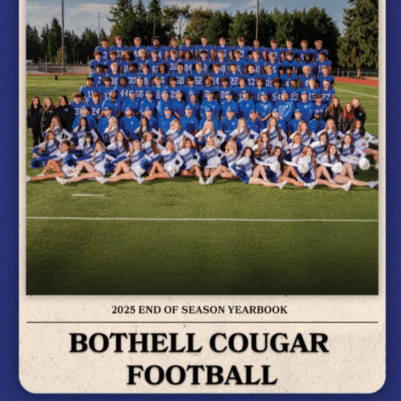 A large group of football players and cheerleaders in blue uniforms pose on a green football field in front of a stadium and trees.