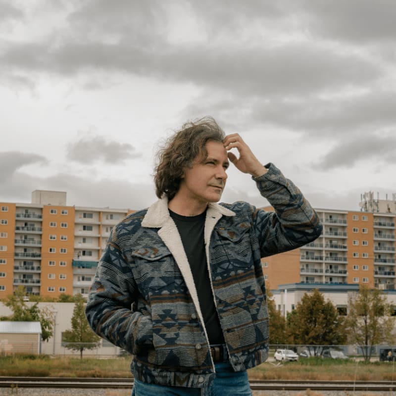 A man in a patterned jacket stands with his hand on his head in front of a tall, orange apartment building under a cloudy sky.