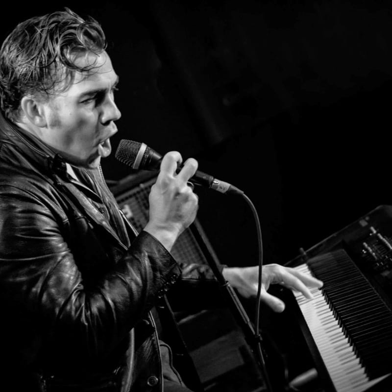 A man in a leather jacket sings into a microphone while playing a keyboard, with a dark, indistinct background.