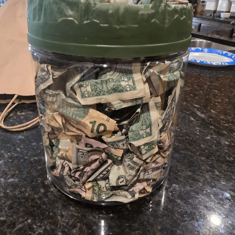 A glass jar filled with crumpled US dollar bills sits on a dark countertop, with a brown paper bag and a blue and white plate visible in the blurred background.