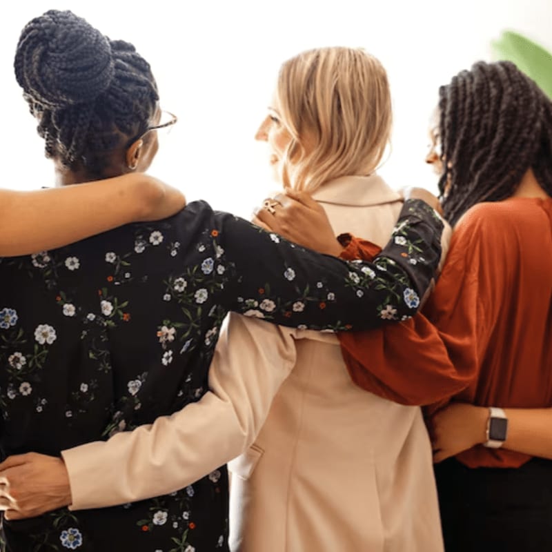 Three women with their arms around each other stand in front of a bright, out-of-focus background.