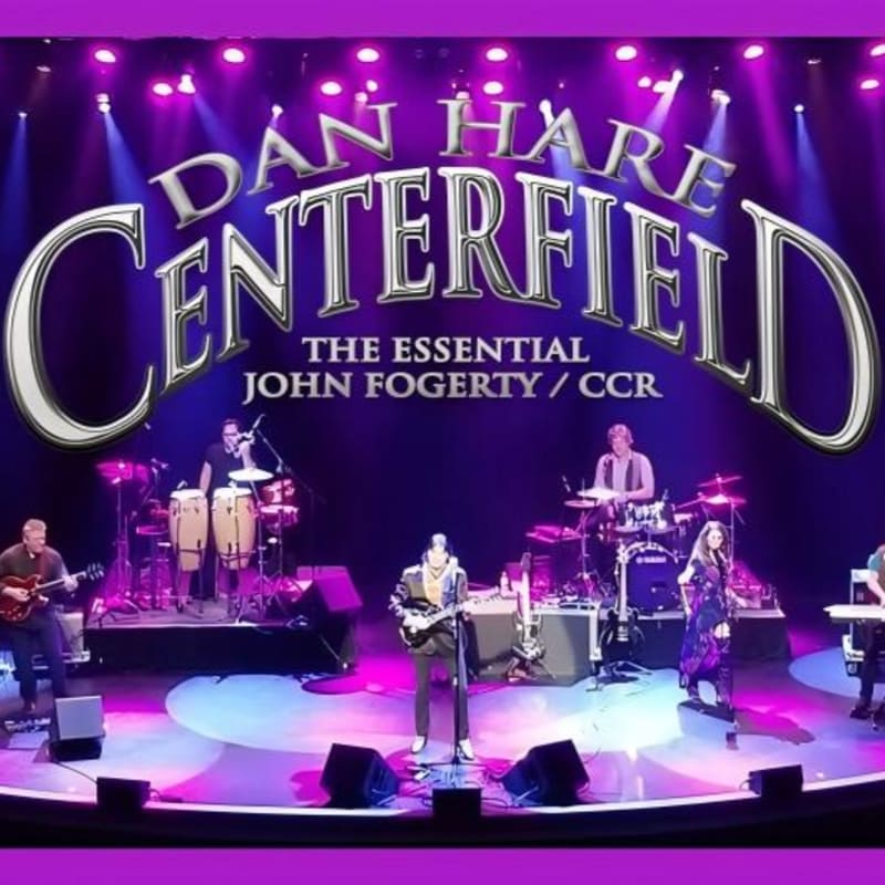 A band performs on a stage with purple lighting and a large "DAN HARE CENTERFIELD" sign in the background, while a keyboardist and guitarist are visible in the foreground.