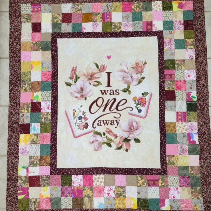 A colorful patchwork quilt with a central panel featuring magnolia blossoms and the words "I was one away" is laid out on a tiled floor.