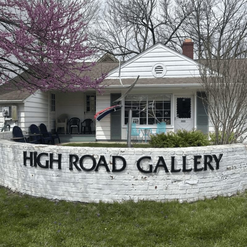 A white brick wall with "HIGH ROAD GALLERY" in black letters curves in the foreground, leading to a white building with a porch and trees in the background.