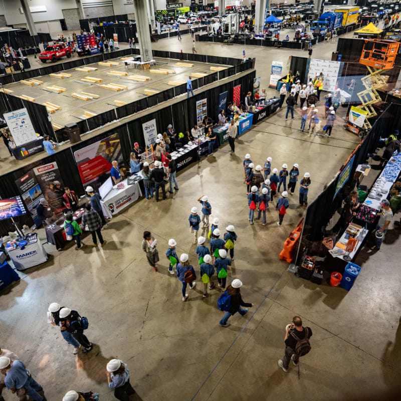 A large indoor exhibition hall is filled with people browsing booths and displays, with a construction-themed exhibit in the foreground and various vehicles and booths visible in the background.