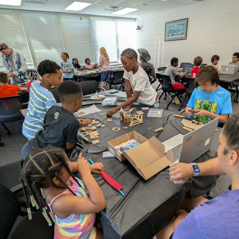 Children and adults are gathered around a table in the foreground, engaged in a craft project, while in the background, more people are working at computers in a brightly lit room.