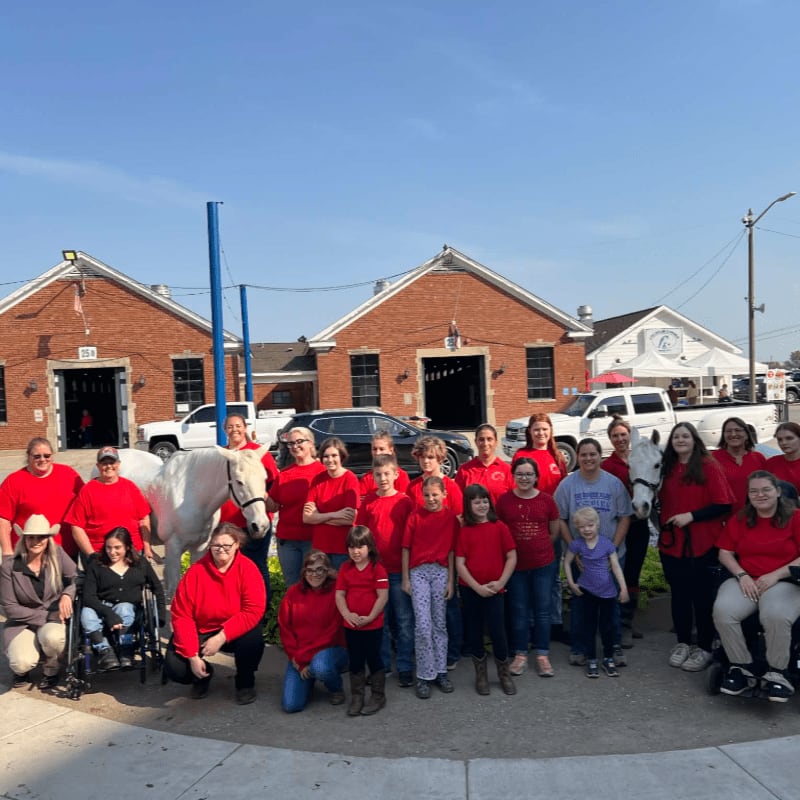 A group of people in red shirts stand with a white horse in front of brick buildings under a clear sky.