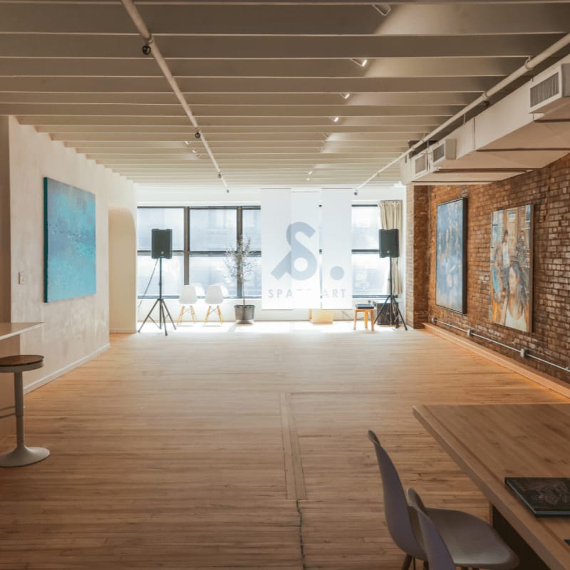 A spacious, light-filled room with a wooden floor and artwork on the walls is visible in the foreground, while large windows and a brick wall form the background.