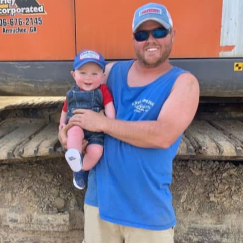 A smiling man in a blue tank top and baseball cap holds a happy baby in overalls in front of a large orange construction vehicle.