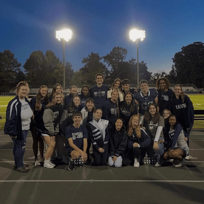 A group of young people in athletic wear pose for a photo on a track in front of illuminated stadium lights and a football field.