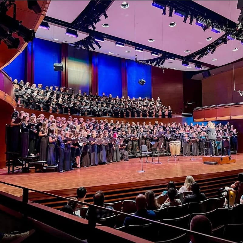 A large choir performs on a brightly lit stage in a concert hall, with an audience seated in the foreground.